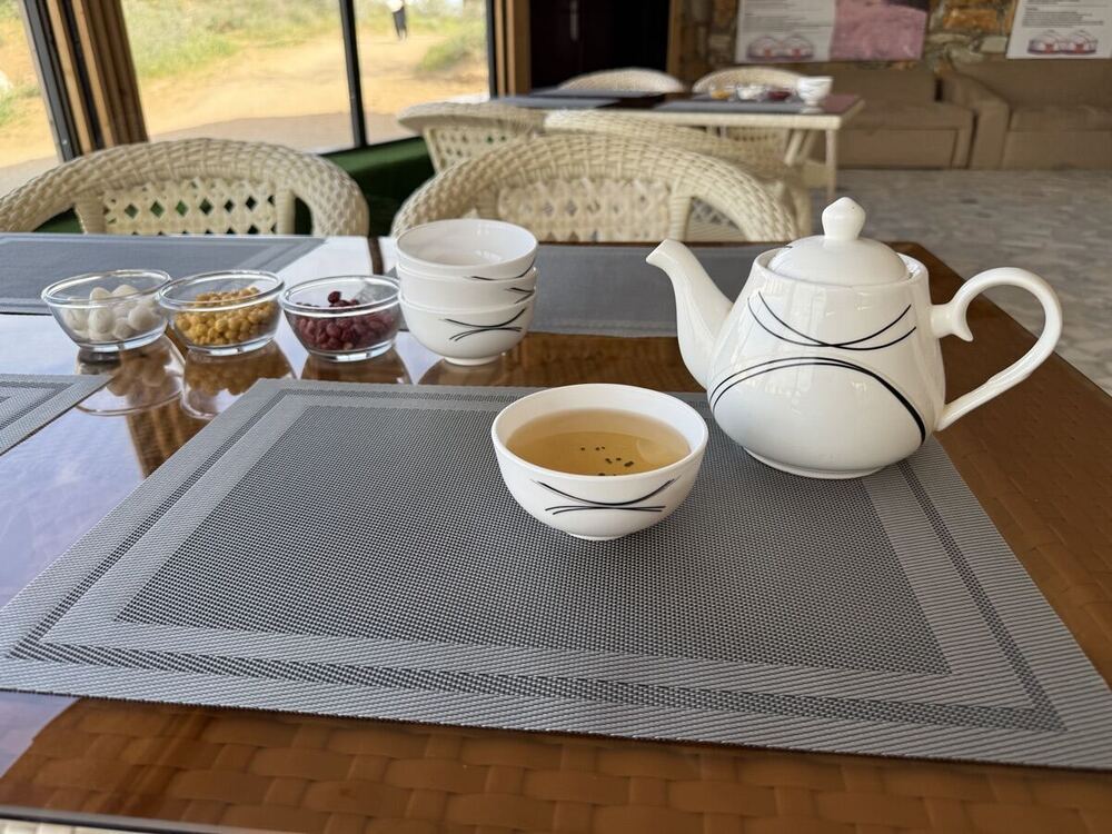 A white teapot, an empty tea bowl with green tea inside, and small dishes of sugar and red snacks on a placemat at the yurt camp common area, with the desert visible through the window in the background.
